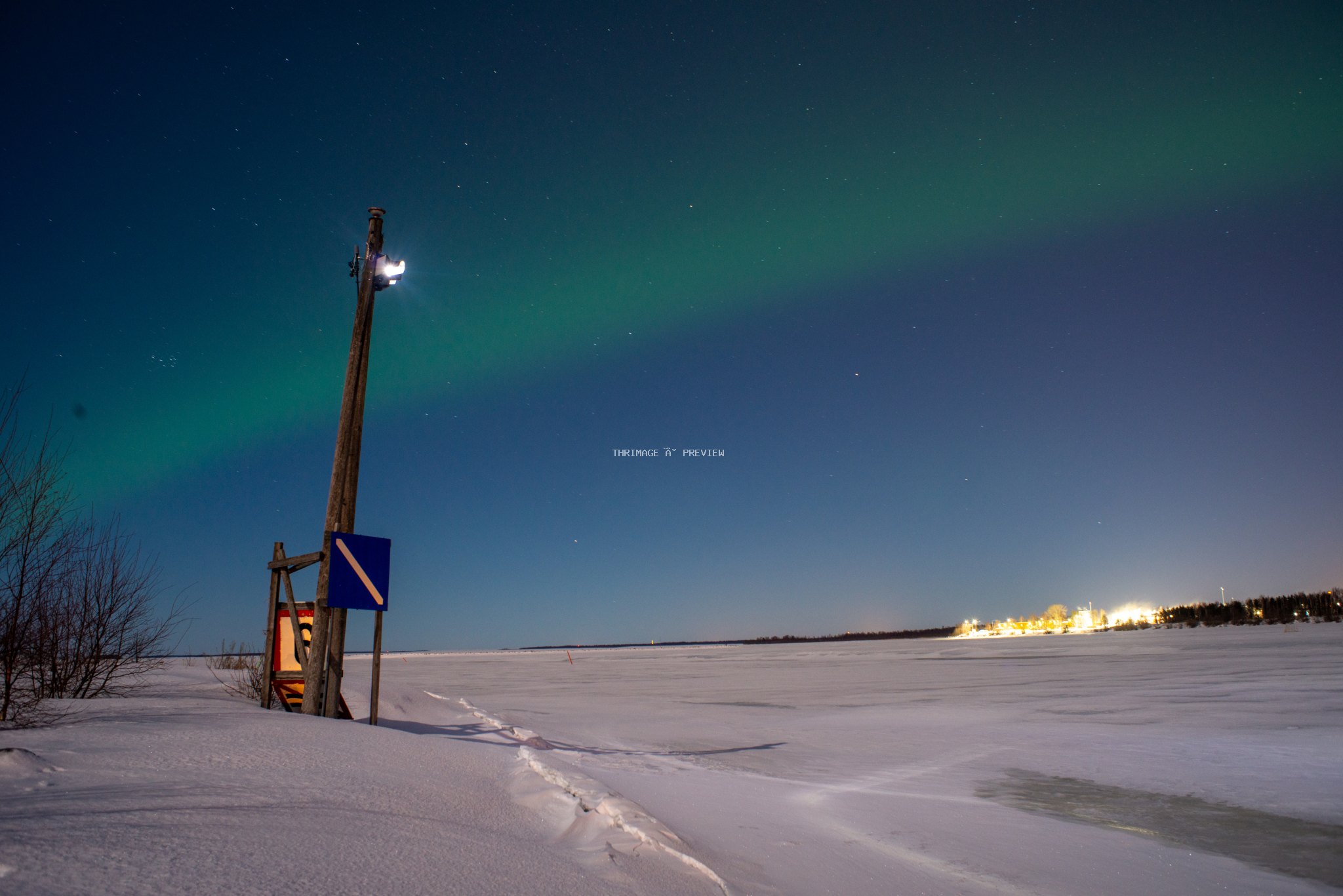 Green aurora glowing above a snowy frozen landscape with a utility pole and distant lights.